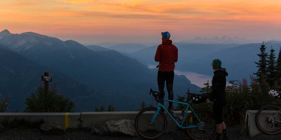 Deux cyclistes gravel arrêtés à un belvédère de montagne au coucher du soleil, l'un portant un manteau de vélo rouge 7mesh