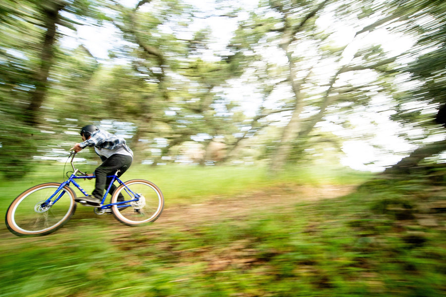 Cycliste sur un Marin Madrone Trail bleu à pneus beiges, filant sur un sentier de terre sous les chênes, avec un effet de mouvement