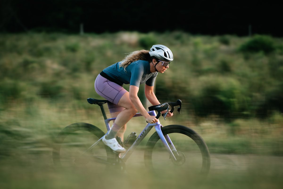 Cycliste en mouvement sur un Scott Addict Gravel violet, sur un chemin de gravelle, devant un talus de végétation floue