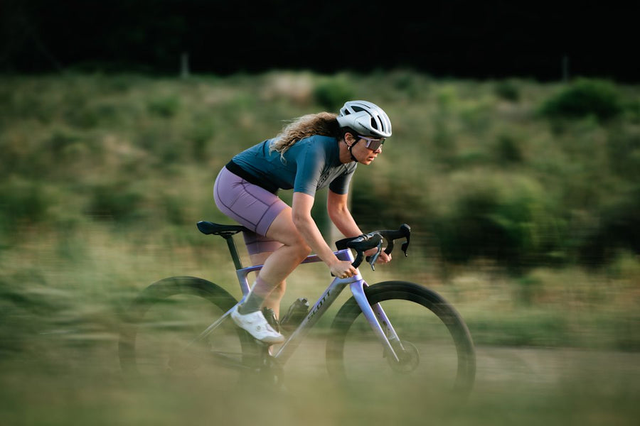 Cycliste en mouvement sur un Scott Addict Gravel violet, sur un chemin de gravelle, devant un talus de végétation floue