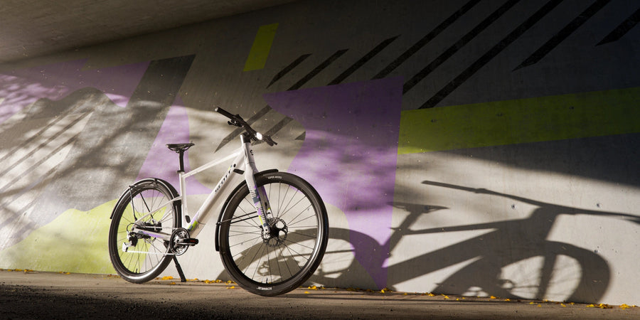 Scott Sweep 10 blanc appuyé contre un mur de béton peint de formes lilas et citron, sous un pont, avec l'ombre du vélo projetée au soleil rasant