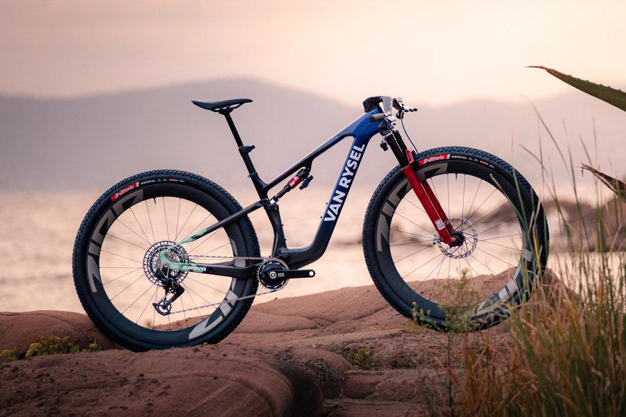 Vélo de montagne Van Rysel RCR-XC à suspension intégrale, cadre carbone bleu et noir, fourche rouge et grandes roues de 32 pouces, posé sur un rocher au bord de la mer au coucher du soleil