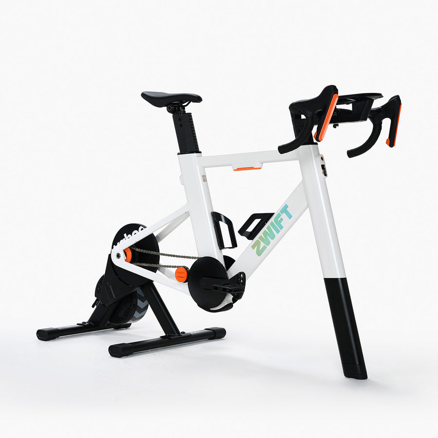 Cadre d'entraînement Zwift Ride V2 blanc monté sur une base Wahoo KICKR CORE 2 noire, vu de trois quarts arrière, avec manettes orange au guidon
